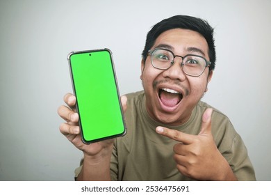  An excited Asian man with glasses holding a smartphone with a green screen, pointing at the device with a wide, enthusiastic expression. - Powered by Shutterstock