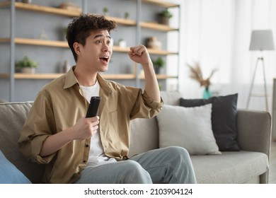 Excited Asian Man Having Fun At Home, Holding Remote Controller In His Hand And Shaking Clenched Fist, Watching Football Game On TV. Happy Guy Enjoying Weekend, Watching Sports Match In Living Room