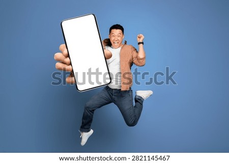 Excited Asian Man Jumping While Holding Smartphone With Blank Screen, Studio Background