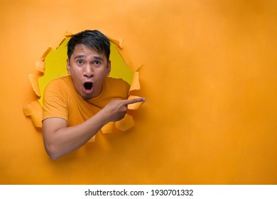 An Excited Asian Man With Shock Face Pointing A Copy Space, Poses On Torn Paper Hole.