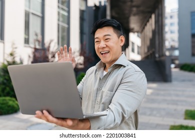 Excited Asian Mature Businessman Having Video Call On Laptop, Waving Hand To Webcamera, Sitting Near Modern Office Building Outdoors. Business Meeting Remote, Coffee Break, Social Distance