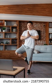 Excited Asian Mature Man In Sportswear Attending Online Yoga Class, Standing In Tree Pose In Front Of Laptop And Smiling, Home Interior, Copy Space, Full Length Shot