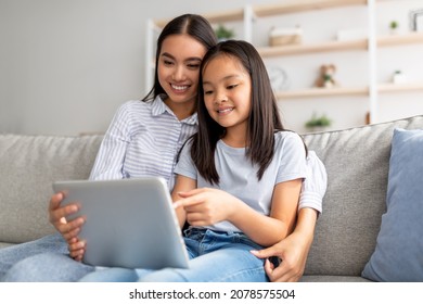 Excited asian mother and daughter using digital tablet together while sitting on sofa and surfing internet at home, free space. Happy mom and child playing online games on pad and hugging