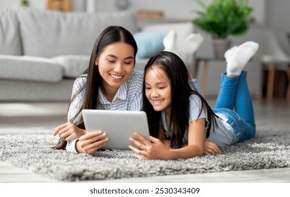 Excited asian mother helping her daughter with lesson on digital tablet, lying together on floor carpet in living room interior. Young mother and her child making online home assignment togeher - Powered by Shutterstock