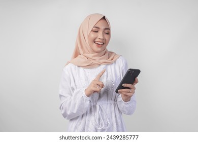 Excited Asian Muslim woman wearing white dress and hijab holding her smartphone while smiling cheerfully, isolated by white background. Ramadhan and Eid Mubarak concept - Powered by Shutterstock