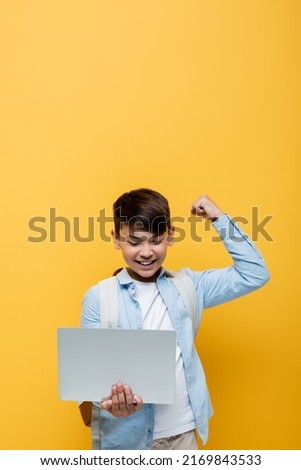 Excited asian schoolkid showing yes gesture and holding laptop isolated on yellow