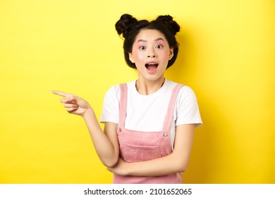 Excited Asian Teen Girl Pointing Finger Left At Logo And Looking Happy, Showing Advertisement, Standing On Yellow Background