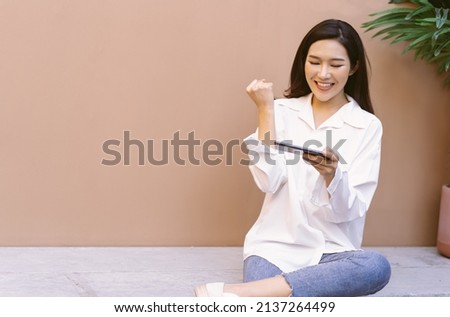 Excited Asian woman celebrating success, showing yes gesture with hands, winner of the competition showing hand fist and looking at mobile phone while sitting on the stone bench in garden at home