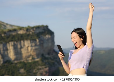 Excited Asian Woman Checking Smart Phone In The Mountain On Vacation