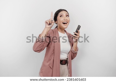 Excited Asian woman employee wearing cardigan, holding her phone, and pointing at the copy space above her, isolated by white background