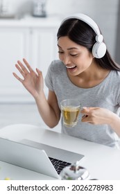 Excited Asian Woman In Headphones Holding Cup Of Tea And Waving Hand During Video Call