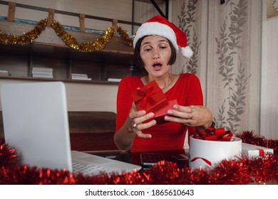 Excited Asian Woman Holding And Opening Christmas Present, Smiling Lady Unwrapping Gift Box, Sitting On The Table At Home In Living Room With Laptop Computer, Celebrating Online