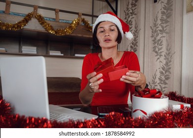 Excited Asian Woman Holding And Opening Christmas Present, Smiling Lady Unwrapping Gift Box, Sitting On The Table At Home In Living Room With Laptop Computer, Celebrating Online