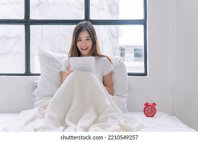 Excited Asian Woman Looking Digital Tablet Good News Working Online On White Bed Beside Window At Home. Happy Young Female Sitting Relax With Tablet Communication Looking Something At Home. Technology