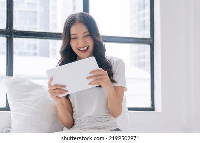 Excited asian woman play gaming online with tablet sitting beside window at home.Joyful young girl holding tablet touchpad playing game application fun relaxed with pillow indoors.Lifestyle technology - Powered by Shutterstock