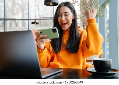 Excited asian woman playing online game on cellphone and making winner gesture in cafe