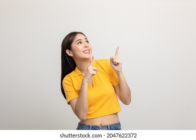 Excited asian woman pointing the finger to blank space over head for advertising text on isolated background. Joyful teenage girl in yellow shirt standing in white room looking at empty space.
