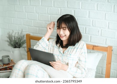 Excited Asian woman with raised hands watching movie on tablet in bedroom - Powered by Shutterstock