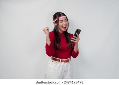 Excited Asian woman in a red shirt holding a cell phone and while raising fist to celebrate victory or a happy moment. Indonesia Independence Day concept. - Powered by Shutterstock