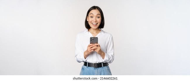 Excited asian woman smiling, reacting to info on mobile phone, holding smartphone and looking happy at camera, standing over white background - Powered by Shutterstock