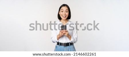 Excited asian woman smiling, reacting to info on mobile phone, holding smartphone and looking happy at camera, standing over white background