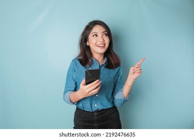 Excited Asian woman wearing blue shirt pointing at the copy space beside her while holding a smartphone, isolated by blue background - Shutterstock ID 2192476635