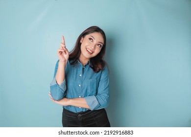 Excited Asian woman wearing blue shirt pointing at the copy space upwards, isolated by blue background