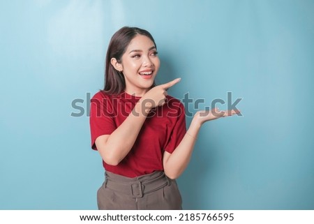 Excited Asian woman wearing red t-shirt pointing at the copy space beside her, isolated by blue background