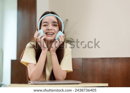 Excited Asian young adult in clear spectacles, sky blue headphones in grasp, expressing joy, workspace around. Elated person holding teal audio gear, shiny orthodontics on display, home setting