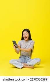 Excited Asian Young Girl Sitting On The Floor Holding Smartphone Texting Or Shopping Online Or Win Gaming Isolated On Yellow Background. Product Placement. Mobile App Advertisement. Copy Space. 