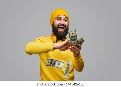 Excited bearded hipster in yellow outfit throwing money in air and looking at camera against gray background