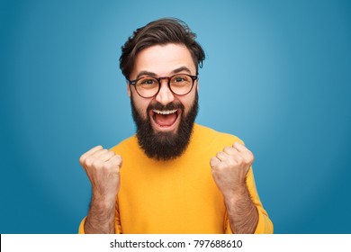 Excited Bearded Man In Glasses Standing With Fists Up On Blue Background. 