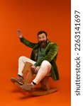 Excited bearded man in jacket waving hand while sitting on rocking horse on red background