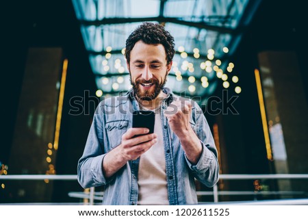 Excited bearded young man blogger cheering favourite team during live stream on website on modern smartphone.Cheerful hipster guy celebrating victory in internet contest on web page holding cellular