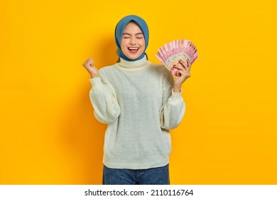 Excited beautiful Asian Muslim woman in white sweater holding cash money in Indonesian rupiah banknotes and celebrating luck isolated over yellow background. People religious lifestyle concept - Shutterstock ID 2110116764