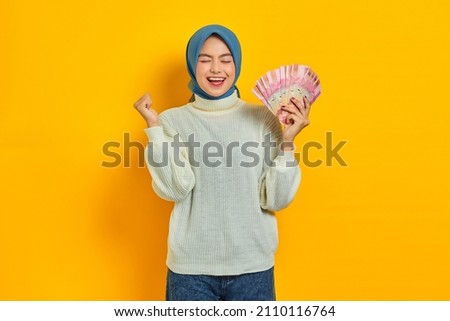 Excited beautiful Asian Muslim woman in white sweater holding cash money in Indonesian rupiah banknotes and celebrating luck isolated over yellow background. People religious lifestyle concept