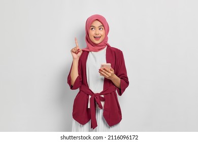 Excited beautiful Asian woman in casual shirt and hijab holding mobile phone and pointing finger up, creating genius solutions isolated over white background. People religious lifestyle concept - Shutterstock ID 2116096172