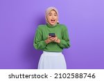 Excited beautiful Asian woman in green sweater and hijab using a mobile phone, received good news isolated over purple background