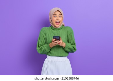 Excited beautiful Asian woman in green sweater and hijab using a mobile phone, received good news isolated over purple background - Powered by Shutterstock