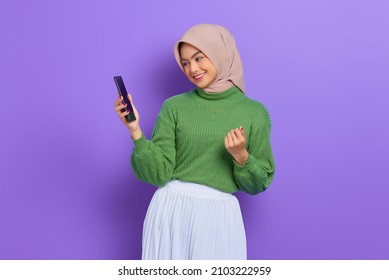 Excited beautiful Asian woman in green sweater and hijab using a mobile phone, celebrating success, getting good news isolated over purple background