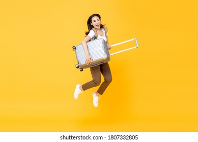 Excited beautiful Asian woman tourist with luggage jumping in mid-air studio shot isolated on yellow background, selective focus