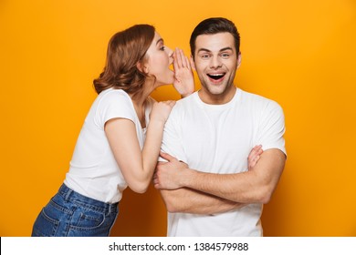 Excited beautiful couple wearing white t-shirts standing isolated over yellow background, telling secret