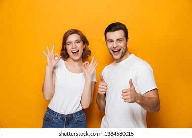 Excited beautiful couple wearing white t-shirts standing isolated over yellow background, showing thumbs up