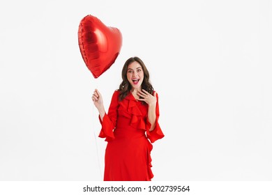 Excited beautiful girl wearing red dress posing with heart balloon isolated over white background