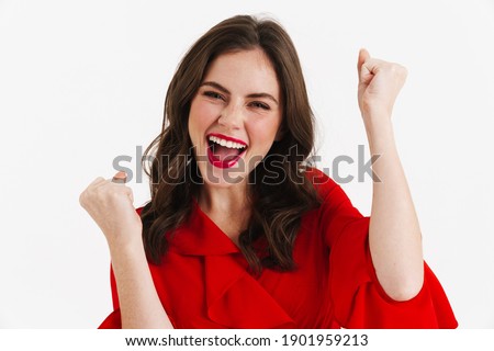 Excited beautiful girl wearing red dress making winner gesture isolated over white background