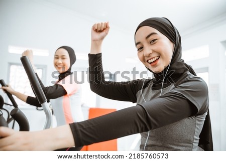 excited beautiful woman with hijab at the gym exercising with friend on static elliptical cycle machine together