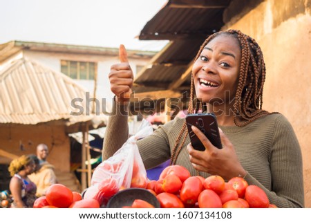 excited beautiful young african woman in a local african market using her smartphone giving thumbs up