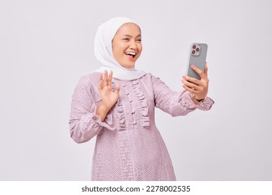 Excited beautiful young Asian Muslim woman wearing hijab and purple dress using smartphone making video call or taking selfie isolated on white studio background - Powered by Shutterstock