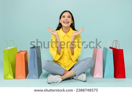 Excited beautiful young lady shopaholic sitting on floor among colorful paper shopping bags, gesturing and smiling, enjoying sale season, black friday deal, isolated on blue background
