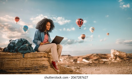 Excited Beautiful Young Multiethnic Female Traveller with Afro Hairstyle Using Laptop Computer on Top of Rocky Canyon Valley. Hot Air Balloons Fly in Mountain National Park. Hiking in Nature. - Powered by Shutterstock